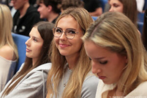 Trzy uczennice (środkowa uśmiechnięta, patrząca w obiektyw) siedzące w auli uniwersyteckiej podczas wydarzenia Szczeciński Salon Maturzystów – Perspektywy 2025. W tle inni uczestnicy spotkania.