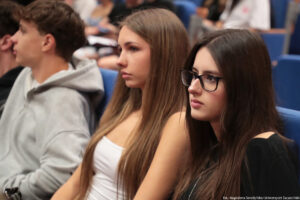 Ujęcie bokiem. Dwie uczennice siedzące w auli uniwersyteckiej podczas wydarzenia Szczeciński Salon Maturzystów – Perspektywy 2025. W tle inni uczestnicy spotkania.