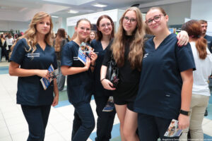 Kilka kobiet ubranych w granatowe kitle i trzymających ulotki z ofertą swojej uczeni podczas wydarzenia Szczeciński Salon Maturzystów – Perspektywy 2025. W tle stoiska szkół wyższych i uczestnicy spotkania.