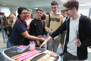 Bokiem po lewej uśmiechnięta kobieta zakładająca na rękę opaskę odblaskową uczniowi stojącemu po lewej. W środku dwóch innych uczniów. Na stoliku obok kobiety informator, długopisy w kubku, odblaski i zakładki z kolorowymi karteczkami.