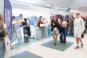 W tle stoiska szkół wyższych i stojących przy nich uczestnicy spotkania. Z przodu przechodzący korytarzem uczniowie (dwie dziewczyny i jeden chłopak).