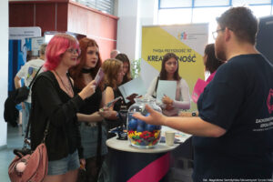 Po prawej bokiem mężczyzna obsługujący stoisko, na którym leżą informatory z ofertą edukacyjną i gadżety. Po lewej zainteresowane uczennice trzymające informatory. W te rollup z napisem na żółtym tle: Twoja przyszłość to kreatywność.