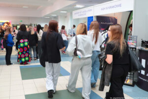 Po prawej stoiska szkół wyższych i stojących przy nich uczestnicy Szczecińskiego Salonu Maturzystów. Po lewej przechodzący korytarzem uczniowie (wszyscy tyłem).