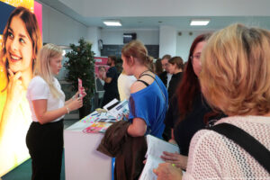 Po prawej bokiem kobieta prezentująca ulotkę i obsługująca stoisko, na którym leżą informatory i gadżety. Po lewej zainteresowane ofertą edukacyjną uczennice. W tle ścianka z uśmiechniętą studentką.