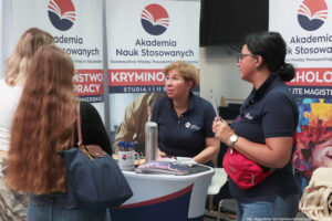 Przodem po prawej dwie kobiety prezentujące ofertę edukacyjną. Tyłem po lewej zainteresowane uczennice. Na stoliku informatory, długopisy w kubku i inne gadżety. W tle rollupy z logiem Akademii Nauk Stosowanych.