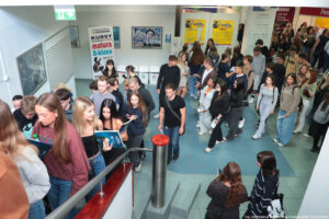 Około 50-osoba grupa uczniów wchodzi po schodach w budynku Uniwersytetu Szczecińskiego. W tle żółte plakaty z napisem Salon Maturzystów i programem wydarzenia.