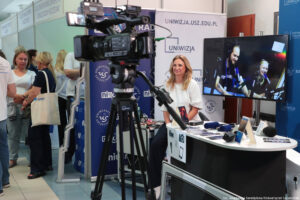 Na środku tyłem kamera na statywie. Przed kamerą uśmiechnięta dziewczyna. W tle rollupy z logiem Uniwizji i NiUS Radia. Po prawej stronie telewizor i stolik z mikrofonami, po lewej stoiska uczelni i uczestnicy Szczecińskiego Salonu Maturzystów.