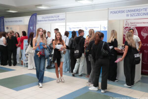 W tle stoiska szkół wyższych i stojących przy nich uczestnicy spotkania. Z przodu przechodzące korytarzem dwie uczennice trzymające informatory.