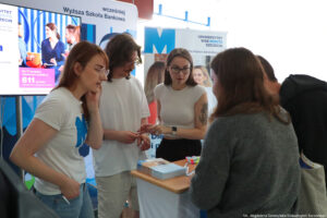 Po prawej bokiem dwie kobiety i mężczyzna prezentujący ofertę edukacyjną swojej uczelni. Po lewej zainteresowani uczniowie (dziewczyna i chłopak). W tle ścianki i rollupy z logiem WSB Merito.