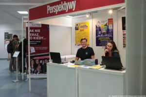 Stoisko. U góry biały napis Perspektywy na czerwony tle. Na stoisku kobieta i mężczyzna, obsługujący laptopy, stojące obok informatorów. W tle plakaty dotyczące Szczecińskiego Salonu Maturzystów.