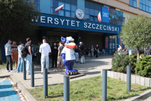 Ujęcie bokiem. Budynek z dużym białym napisem Uniwersytet Szczeciński. Nad nim zawieszone dwie biało-czerwone flagi. Przed budynkiem uczniowie i figura około 2-metrowego gryfusia z koszulką z logiem uczelni.