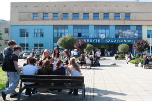 Budynek z dużym białym napisem Uniwersytet Szczeciński. Nad nim zawieszone dwie biało-czerwone flagi. Przed budynkiem uczniowie – stojący i siedzący na ławkach ustawionych obok nasadzeń.