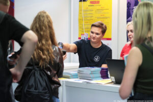 Stoisko Perspektyw. Przodem mężczyzna skanujący kod w telefonie stojącej tyłem uczestniczki Szczecińskiego Salonu Maturzystów. Za nim na żółtym plakacie program wydarzenia. Przed nim na stoliku – informatory i laptop.