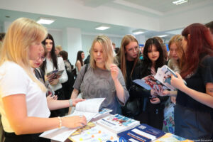 Po lewej bokiem kobieta obsługująca stoisko, na którym leżą informatory i ulotki. Po prawej zainteresowane uczennice przeglądające informatory Uniwersytetu Szczecińskiego.