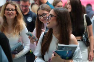 Trzy uśmiechnięte uczennice trzymające informatory podczas Szczecińskiego Salonu Maturzystów – dwie przodem, jedna bokiem. W tle inni uczestnicy wydarzenia.