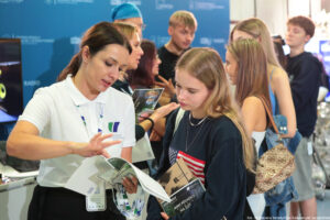 Po lewej bokiem kobieta prezentująca ofertę edukacyjną swojej uczelni. Po prawej zainteresowana uczennica. W tle inni uczestnicy wydarzenia.