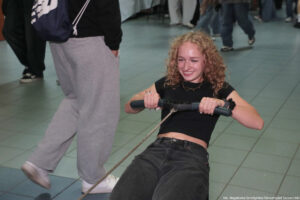 Siedząca, uśmiechnięta uczennica trzymająca uchwyt na ergometrze wioślarskim. Ujęcie do kolan.