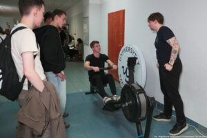 Siedzący, uśmiechnięty uczeń trzymający uchwyt na ergometrze wioślarskim. Po lewej inni uczniowie, po prawej mężczyzna z obsługi.