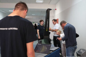 Na środku ergometr narciarski i uczennica próbująca swoich sił na tym urządzeniu. Dookoła niej obsługa.