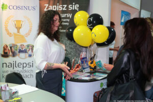 Po lewej bokiem kobieta prezentująca ofertę edukacyjną swojej uczelni. Po prawej zainteresowana uczennica. Na stoliku ulotki i wazon z czarnymi i żółtymi balonami.