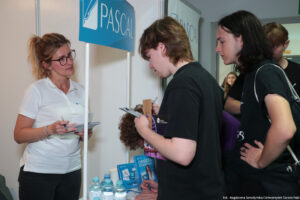 Po lewej bokiem kobieta prezentująca ofertę edukacyjną swojej uczelni. Po prawej trzej zainteresowani uczniowie. Na stoliku ulotki i wody w butelkach.