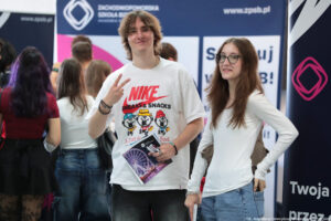 Uśmiechnięci uczniowie (chłopak po lewej, dziewczyna po prawej) podczas Szczecińskiego Salonu Maturzystów. Chłopak trzyma jedną ręką informator i długopisy, drugą pokazuje znak Victorii skierowany zamkniętą dłonią w swoją stronę. W tle tyłem inni uczestnicy wydarzenia.