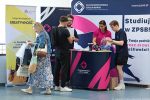 Przedstawiciele Zachodniopomorskiej Szkoły Biznesu (dwóch mężczyzn i jedna kobieta) przedstawiają uczniowi (stoi tyłem) ofertę edukacyjną. Na stoliku przed nimi stoją gadżety, m.in. długopisy i piłeczki. W tle granatowo-biało-różowa ścianka z logiem uczelni.