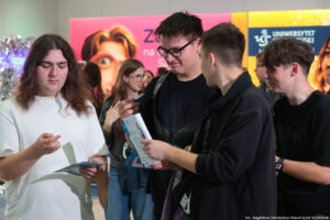 Czterech uśmiechniętych uczniów ogląda długopisy, które otrzymali na jednym ze stoisk podczas Szczecińskiego Salonu Maturzystów. W tle inni uczestnicy wydarzenia i ścianki Uniwersytetu Szczecińskiego – różowa i żółta.