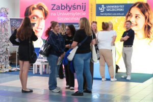 Kilka uczennic stoi na korytarzu podczas Szczecińskiego Salonu Maturzystów. W tle stoisko Uniwersytetu Szczecińskiego i ścianki – różowa z hasłem: Zabłyśnij na Uniwersytecie Szczecińskim i żółta z uśmiechniętą studentką, poziomym logiem US i adresem strony: kandydaci.usz.edu.pl.