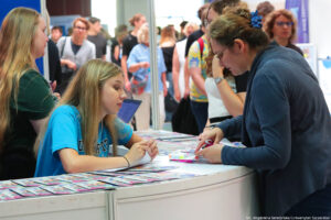 Po lewej bokiem kobieta prezentująca ofertę edukacyjną swojej uczelni. Po prawej osoby zainteresowane. Na stoliku ulotki.