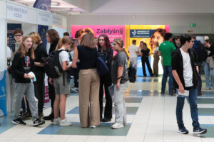 Kilkunastu uczniów stoi na korytarzu i przy stoiskach uczelni podczas Szczecińskiego Salonu Maturzystów. W tle stoisko Uniwersytetu Szczecińskiego i ścianki – różowa z hasłem: Zabłyśnij na Uniwersytecie Szczecińskim i żółta z uśmiechniętą studentką, poziomym logiem US i adresem strony: kandydaci.usz.edu.pl.