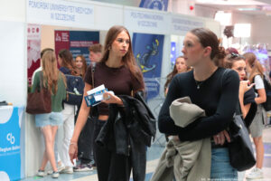 Dwie uczennice trzymające kurtki. Dziewczyna po lewej stronie w jednej dłoni trzyma ulotki. W tle stoiska szczecińskich uczelni i uczestnicy Szczecińskiego Salonu Maturzystów.