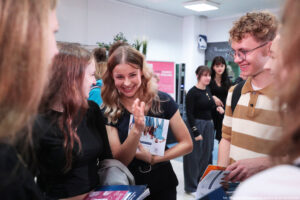Grupa młodych osób rozmawiających w jasnym, wewnętrznym pomieszczeniu. Na pierwszym planie, pośrodku, stoi uśmiechnięta kobieta o jasnej skórze i długich, falowanych blond włosach; ma na sobie czarną sukienkę i trzyma ulotkę lub broszurę z nadrukowanymi postaciami. Po lewej stronie obok niej jest kobieta o jasnej skórze i długich, ciemnorudych włosach w czarnej bluzce, zwrócona w jej stronę, jak w trakcie rozmowy. Po prawej stoi mężczyzna o jasnej skórze, kręconych jasnobrązowych włosach i w okularach; ma koszulkę w szerokie beżowo-białe pasy i plecak na ramieniu, trzyma kilka broszur lub kartek. Na dole kadru leżą stosy kolorowych materiałów papierowych (broszury/ulotki). W tle widać kilka kolejnych osób, w tym dwie kobiety w ciemnych ubraniach, oraz elementy wnętrza: białe ściany, oświetlenie sufitowe i tablice/plakaty; część napisów jest nieczytelna z tej odległości. W prawym dolnym rogu znajduje się podpis: „fot. Magdalena Seredyńska/Uniwersytet Szczeciński”.