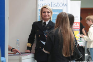 Kobiet w ciemnogranatowym mundurze ze złotymi elementami trzyma ulotkę swojej uczelni i rozmawia ze stojącą tyłem do kadru uczennicą. Na stoliku po lewej stronie ulotki i informatory. Po prawej w tle stoiska innych uczelni i inni uczestnicy wydarzenia.