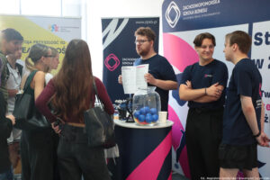 Przedstawiciele Zachodniopomorskiej Szkoły Biznesu (trzech mężczyzn) na stoisku swojej uczelni. Pierwszy z lewej trzyma rozłożony informator i przedstawia ofertę edukacyjną uczniom (stojące bokiem trzy dziewczyny i dwóch chłopaków za nimi). Na stoliku stoją gadżety, m.in. długopisy i piłeczki w słoju. W tle granatowo-biało-różowa ścianka i rollup z logiem uczelni.