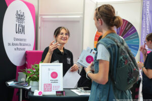 Po lewej stojąca przodem uśmiechnięta kobieta prezentuje ofertę edukacyjną swojej uczelni. Za nią granatowo-biało-różowa ścianka z logiem Uniwersytetu Adama Mickiewicza w Poznaniu. Na stoliku z przodu ramka z QR kodem do sprawdzenia kierunków studiów. Po prawej stojąca tyłem do kadru zainteresowana uczennica. Obok niej kolorowe koło fortuny UAM.