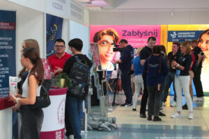 Po lewej stoiska uczelni i stojący przy nich zainteresowani uczniowie. Na środku na końcu operator patrzący w kamerę ustawioną na statywie. Obok kilkuosobowa grupa uczestników Szczecińskiego Salonu Maturzystów. W tle ścianki Uniwersytetu Szczecińskiego – różowa z hasłem: Zabłyśnij na Uniwersytecie Szczecińskim i studentem z lupą oraz żółta z uśmiechniętą studentką, poziomym logiem US i adresem strony: kandydaci.usz.edu.pl.