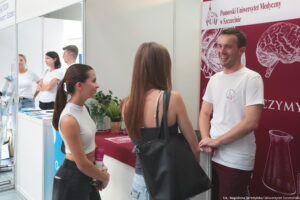 Po prawej uśmiechnięty reprezentant Pomorskiego Uniwersytetu Medycznego opowiadający dwóm uczennicom o ofercie edukacyjnej podczas Szczecińskiego Salonu Maturzystów. Za nim bordowo-biała ścianka z logiem uczelni i mózgiem. W tle (po lewej) stoisko innych uczelni.