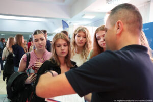 Stojący tyłem mężczyzna trzymający informator i prezentujący ofertę edukacyjną Uniwersytetu Szczecińskiego. Przodem cztery zainteresowane uczennice.