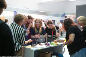 Stoisko uczelni podczas Szczecińskiego Salonu Maturzystów. Na stoliku gadżety i ulotki. Po prawej dwie kobiety obsługujące stoisko (jedna tyłem). Przodem osoby zainteresowane ofertą edukacyjną (kobieta, mężczyzna i trzy uczennice). W tle stoiska innych uczelni.