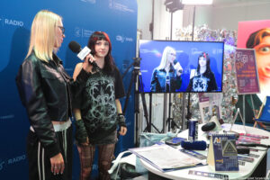 Po lewej stojąca bokiem granatowa ścianka z niebieskimi logami NiUS Radia i Uniwizji. Przed nią dwie dziewczyny – stojąca bokiem dziewczyna po lewej trzyma mikrofon. Obok nich stoi statyw kamery i ekran telewizyjny, na którym obie dziewczyny są widoczne. Przed nim stoi stół z mikrofonami.