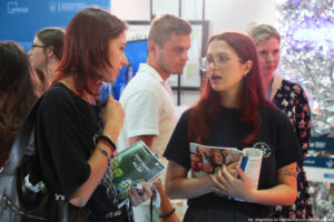 Dwie osoby trzymające informatory i ulotki dyskutują podczas Szczecińskiego Salonu Maturzystów (ujęcie do pasa). W tle inni uczestnicy wydarzenia.