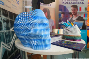 Wydrukowana w drukarce 3D ażurowe czaszki – ta z przodu błękitno-biała, z tyłu biała z elementem różnokolorowym u dołu i włożonymi w nią długopisami. Na stoliku pośrodku leży informator z ofertą edukacyjną. W tle rollup z nazwą uczelni: Politechnika Koszalińska.