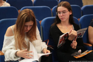 Aula uniwersytecka. Ujęcie bokiem. Na niebieskich fotelach siedzą dwie uczennice i przeglądają informator Uniwersytetu Szczecińskiego podczas Szczecińskiego Salonu Maturzystów – Perspektywy 2025.