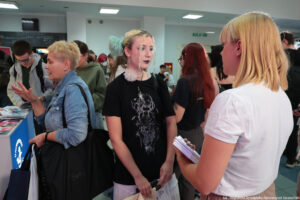 Stojąca tyłem kobieta trzymająca informatory i prezentująca ofertę edukacyjną Uniwersytetu Szczecińskiego. Przodem zainteresowana uczennica. Po lewej stronie fragment stoiska uczelni.