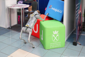 Robot w kształcie psa wspinający się po kostkach – zielonej z białym logiem Uniwersytetu Jagiellońskiego w Krakowie, czerwonej z białą literą U i niebieskiej z napisem: #misjaUJ. W tle stolik z ulotkami.