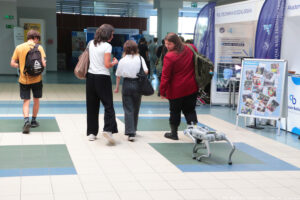 Robot w kształcie psa podąża za trzema uczennicami na korytarzu podczas Szczecińskiego Salonu Maturzystów. Dziewczyna po prawej ogląda się za nim. Mijają stoiska różnych uczelni.