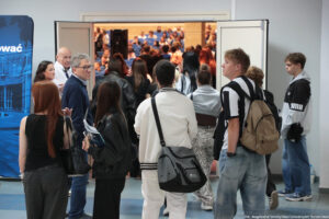 Uczniowie z nauczycielem wchodzący do auli uniwersyteckiej podczas Szczecińskiego Salonu Maturzystów. Większość osób tyłem. W środku auli zgromadzona już spora liczba uczestników siedzących na niebieskich fotelach.