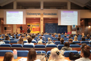 Duża aula uniwersytecka. Ujęcie na scenę. Na niebieskich fotelach siedzą uczniowie (tyłem). Nad sceną ekrany z informacją o egzaminach maturalnych. Na po lewej prelegent, a obok żółta ścianka z napisem: Salon Maturzystów – Perspektywy i element graficzny – studentka z lupą, ubrana w togę i biret.