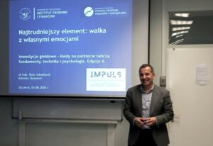 Mężczyzna w garniturze stojący obok ekranu z prezentacją w sali wykładowej. Na ekranie widoczny jest niebieski slajd z białym tekstem w języku polskim. U góry po lewej stronie slajdu znajdują się logotypy i napisy: „UNIWERSYTET SZCZECIŃSKI INSTYTUT EKONOMII I FINANSÓW”. Pośrodku górnej krawędzi slajdu jest okrągłe białe logo, obok którego widnieje tekst: „UNIWERSYTET SZCZECIŃSKI WYDZIAŁ EKONOMII, FINANSÓW I ZARZĄDZANIA”. Centralna część slajdu zawiera duży tytuł: „Najtrudniejszy element: walka z własnymi emocjami”. Niżej, mniejszą czcionką: „Inwestycje giełdowe – kiedy na parkiecie tańczą fundamenty, technika i psychologia. Edycja 4.” Jeszcze niżej: „dr hab. Piotr Szkudlarek Katedra Ekonomii” Na dole po lewej: „Szczecin, 03.06.2026 r.” Po prawej stronie slajdu znajduje się białe prostokątne logo z napisem: „IMPULS” a pod nim mniejszym drukiem, nie w pełni czytelnym, tekst zaczynający się od „KOŁO NAUKOWE…”. Obok ekranu stoi uśmiechnięty mężczyzna o jasnej karnacji, w szarej marynarce i jasnej koszuli z zapiętymi guzikami przy szyi. Trzyma mały przedmiot w dłoniach. W tle widoczny jest fragment białej tablicy i drzwi.